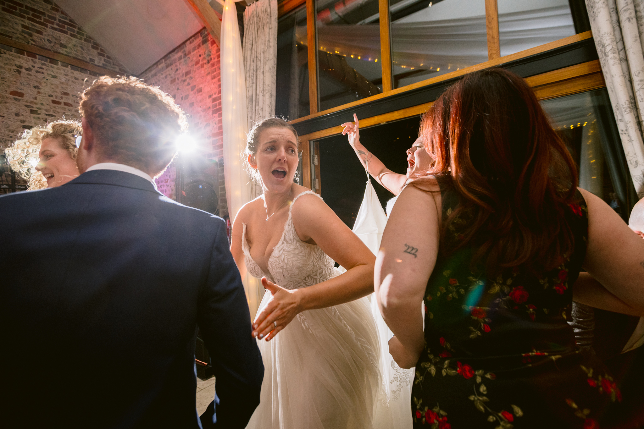 A bride in a white dress dances energetically with guests at a wedding reception, illuminated by bright indoor lighting—capturing the lively spirit of Upwaltham Barns Wedding Photography.