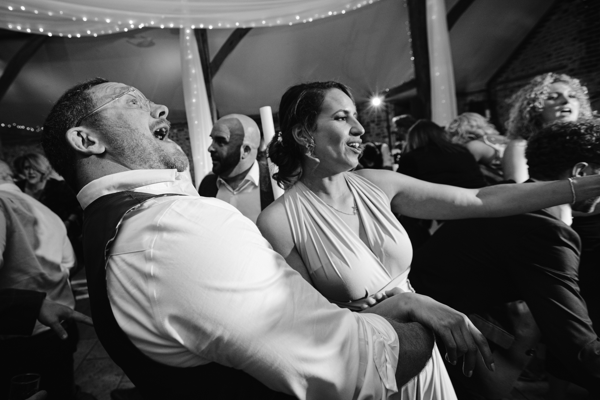 A man and woman dance and smile among a lively group of people at an indoor event with fairy lights overhead, perfectly capturing the joy of Upwaltham Barns Wedding Photography.