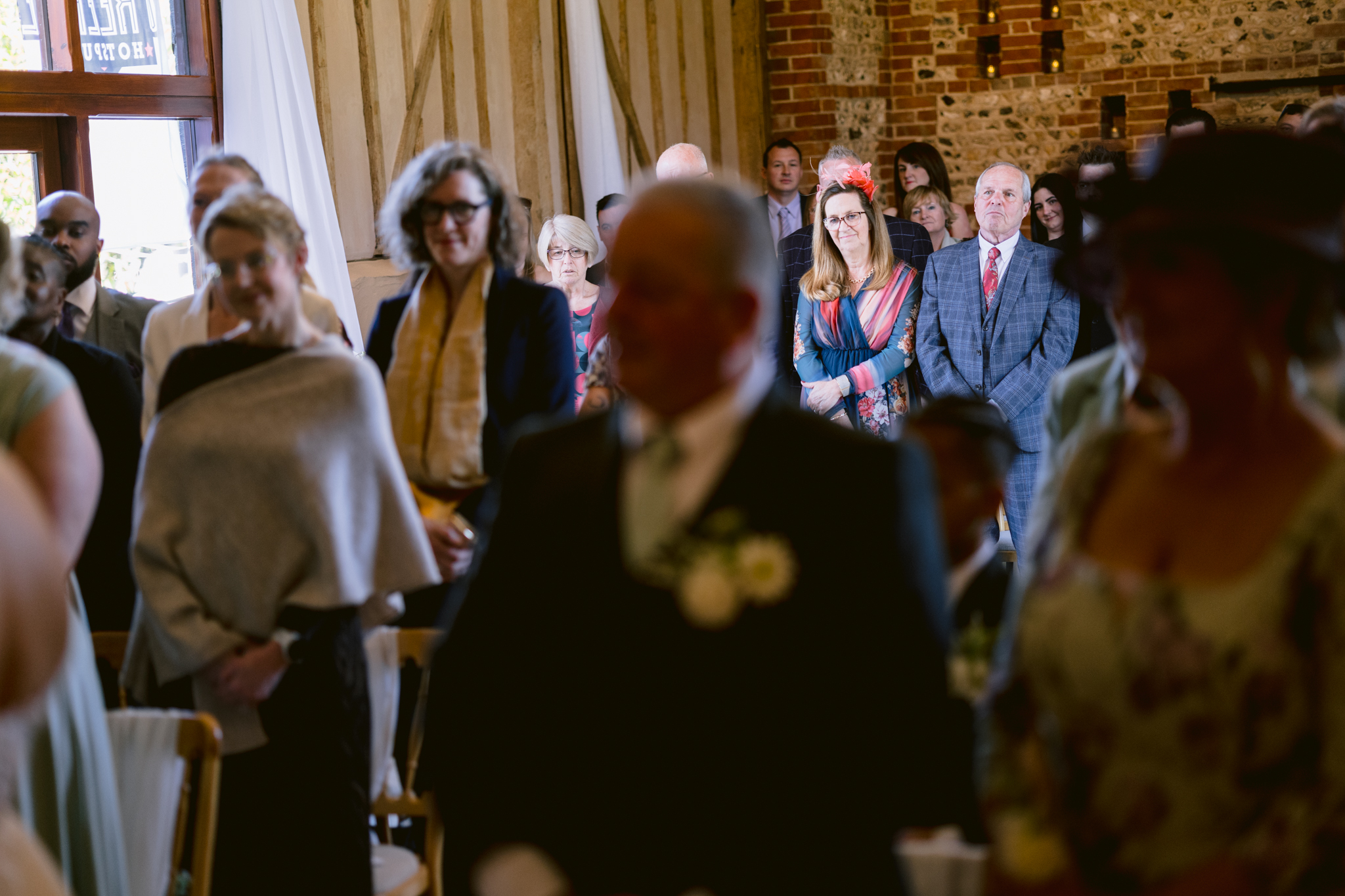 A group of people stand indoors at a formal event, with some seated in the foreground and others watching attentively in the background, capturing the elegant atmosphere typical of Upwaltham Barns Wedding Photography.