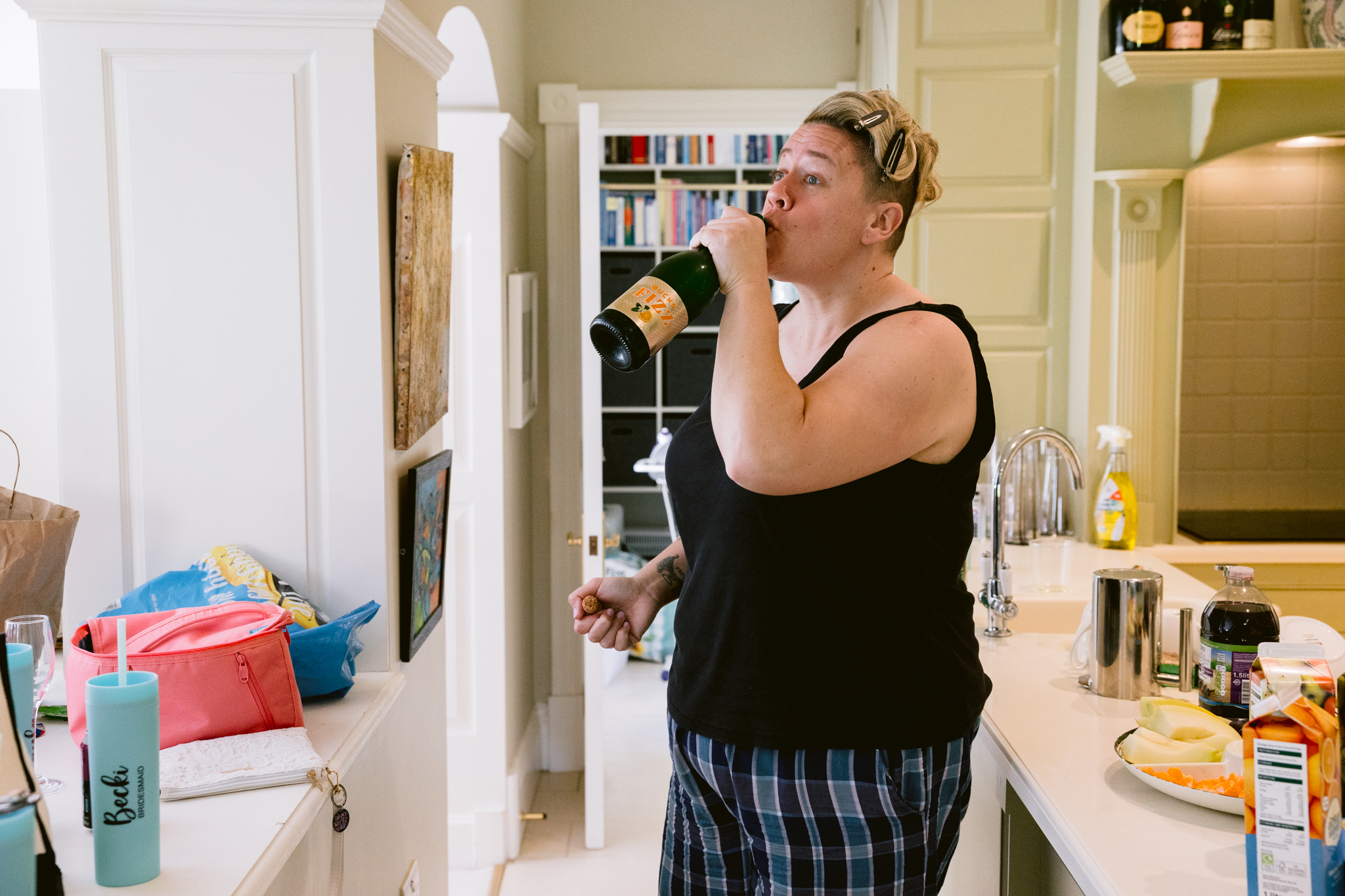 Person in a vest and checked shorts drinks from a bottle in a kitchen with food and drinks on the worktop, capturing a candid moment reminiscent of Upwaltham Barns Wedding Photography style.