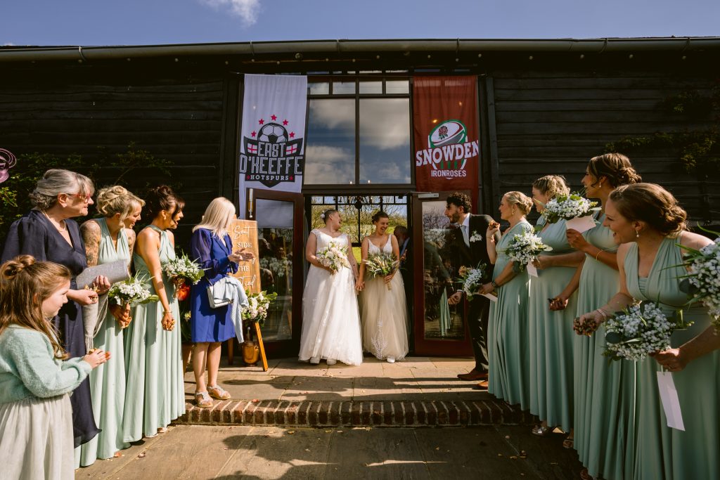 Two brides stand at an entrance holding hands, surrounded by bridesmaids in mint green dresses and guests with bouquets, during a sunny Upwaltham Barns Wedding Photography session.