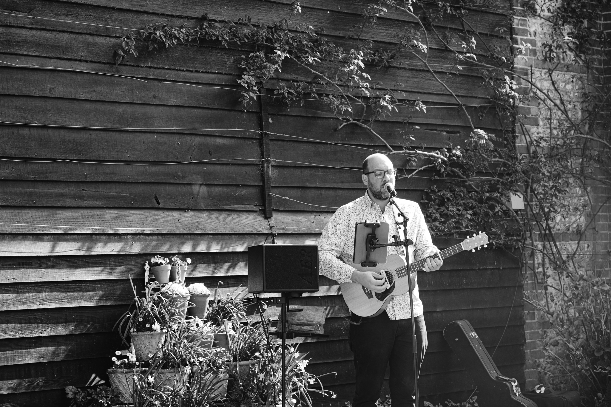 A man plays an acoustic guitar and sings into a microphone outdoors, standing beside a wooden wall with vines and potted plants—an authentic moment captured in Upwaltham Barns Wedding Photography.