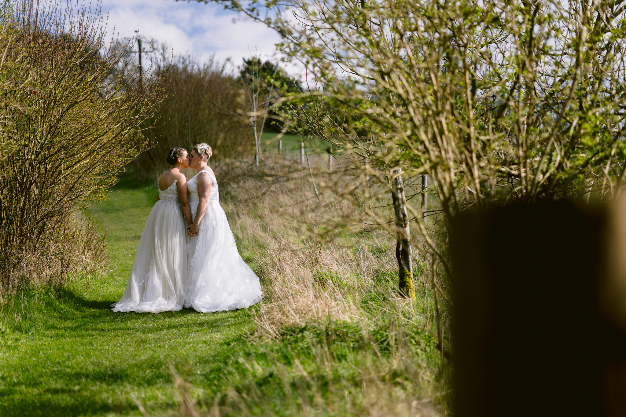 Two women in white wedding dresses stand on a grassy path outdoors at Upwaltham Barns, holding hands and facing each other, surrounded by lush bushes and trees—capturing a beautiful moment of wedding photography.