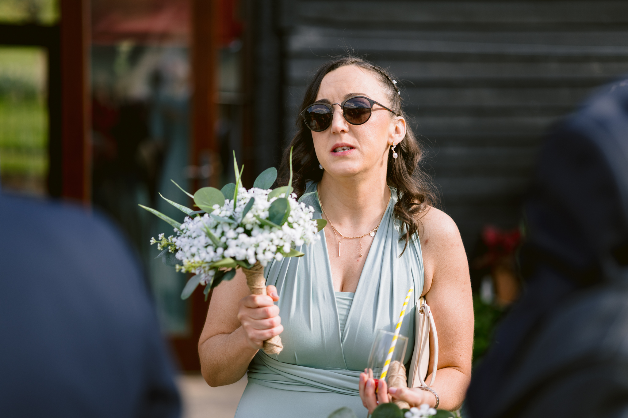 Woman in a light blue dress and sunglasses holds a bouquet of flowers while standing outdoors at Upwaltham Barns, with people and a building in the background—perfect for Upwaltham Barns wedding photography inspiration.