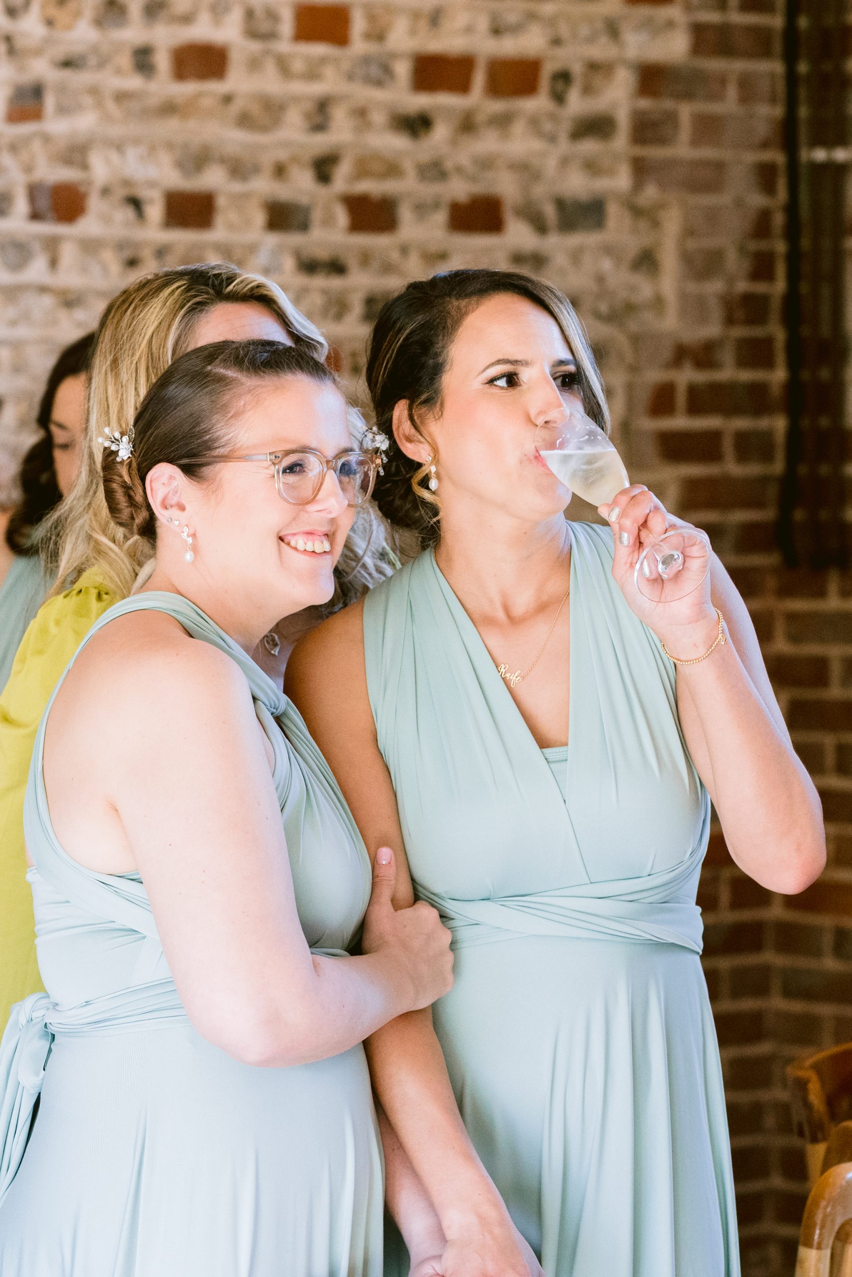 Two women in light blue dresses stand together indoors at an Upwaltham Barns wedding; one smiles at the camera while the other sips from a champagne glass. An exposed brick wall forms the charming backdrop.