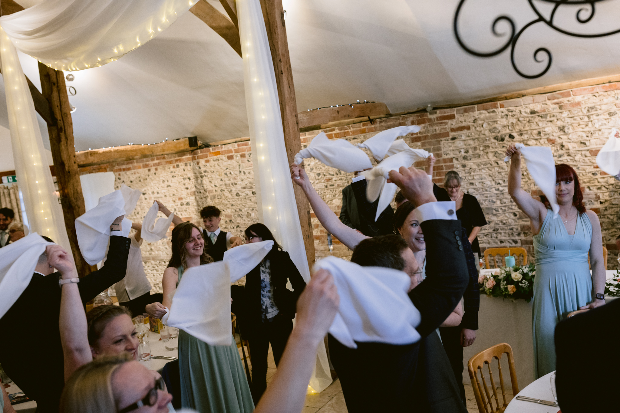 Guests at a formal event wave white serviettes in the air whilst standing around decorated tables in a rustic venue with exposed brick walls and wooden beams—perfectly capturing the charm of Upwaltham Barns Wedding Photography.