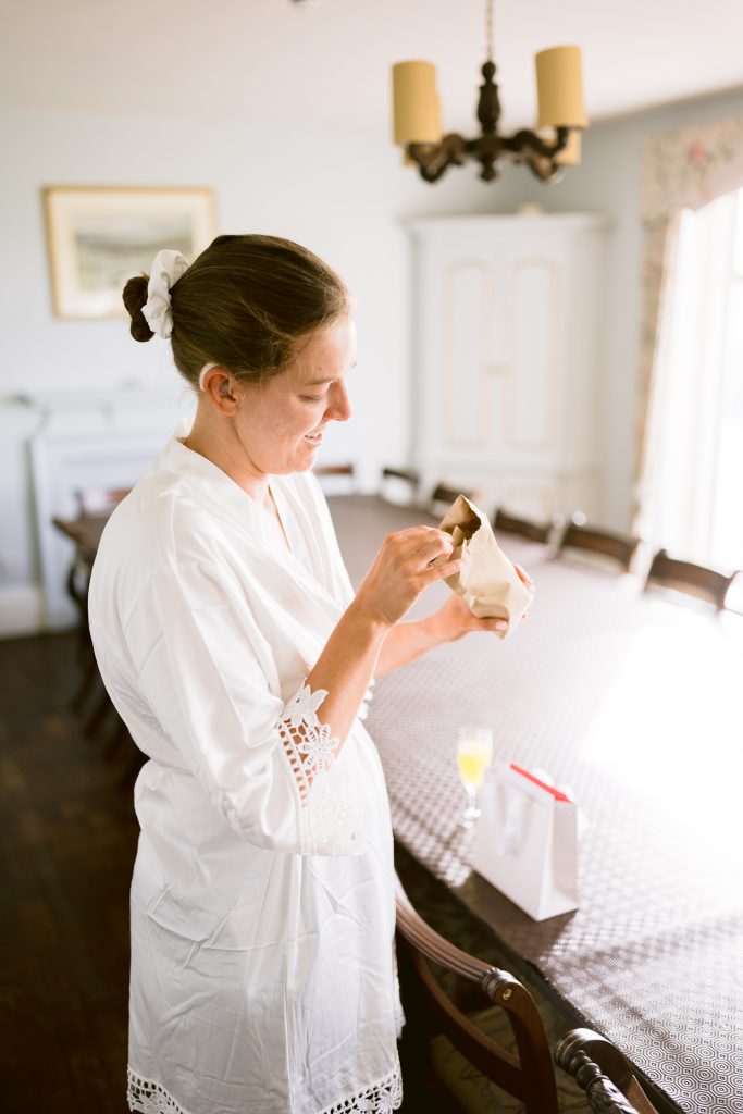 A woman in a white dressing gown stands by a dining table, smiling whilst opening a small gift bag. A glass of orange juice and another bag are on the table—a candid moment perfect for Upwaltham Barns Wedding Photography.