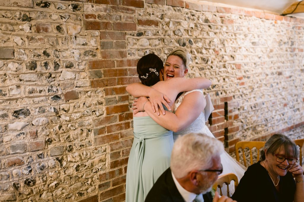 Two women embrace and smile in front of a brick wall at a gathering, whilst three seated people observe and smile nearby—a candid moment captured in the style of Upwaltham Barns Wedding Photography.
