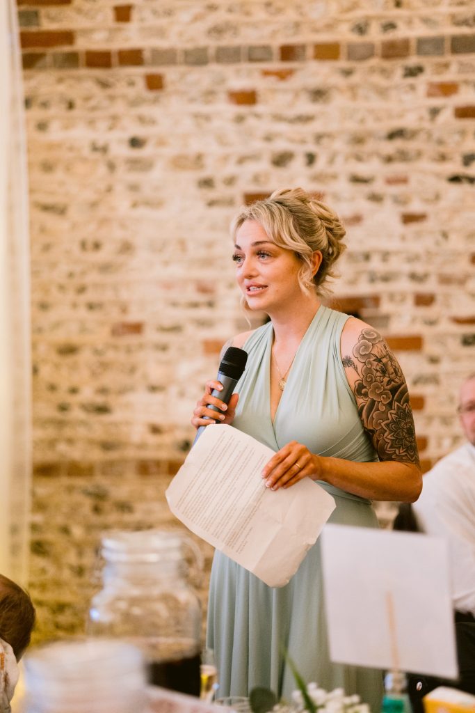 A woman in a light green dress, holding a microphone and a sheet of paper, speaks indoors in front of a brick wall—captured in timeless style by Upwaltham Barns Wedding Photography.