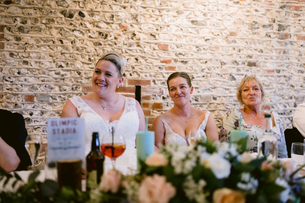 Three women sit at a decorated table, two in white dresses and one in a floral top, all smiling in front of a brick wall—capturing a candid moment perfect for Upwaltham Barns Wedding Photography.