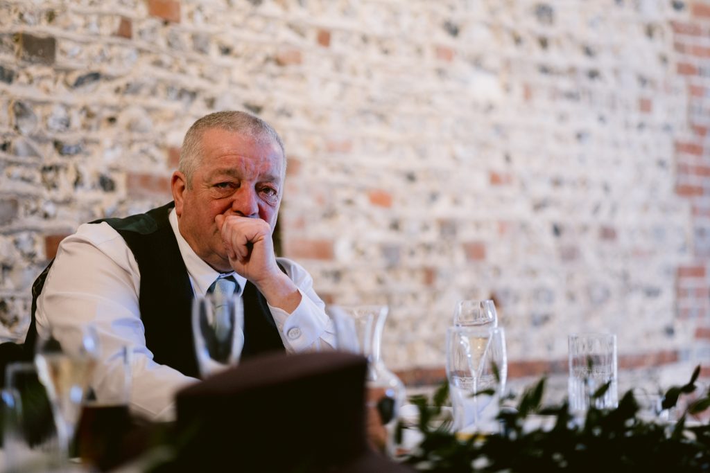 An older man in formal attire sits at a table with empty glasses, resting his chin on his hand, in front of a brick wall—a candid moment captured in Upwaltham Barns Wedding Photography.