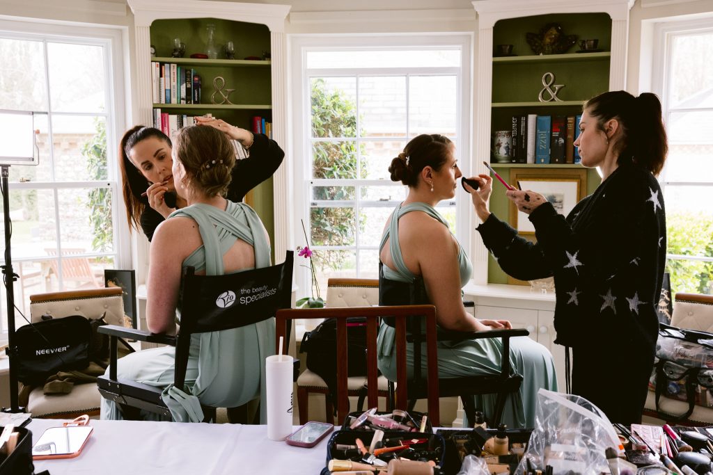 Two women in light blue dresses sit in chairs as two make-up artists apply their make-up, preparing for a special day, in a well-lit room with shelves and large windows—a beautiful moment captured by Upwaltham Barns Wedding Photography.