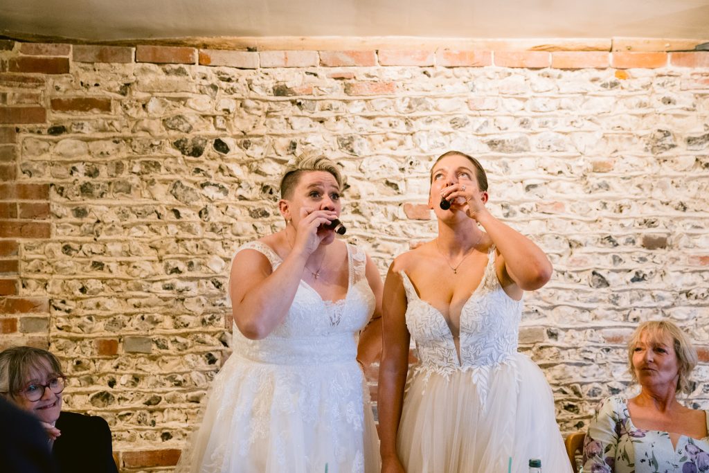 Two women in wedding dresses stand side by side, drinking from small bottles at a table, as two seated women look on—all set against a charming brick and stone wall, beautifully captured in this Upwaltham Barns Wedding Photography moment.