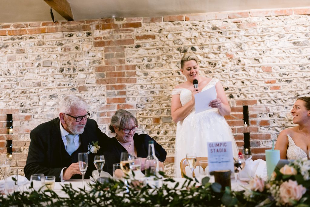 A woman in a white dress gives a speech at an Upwaltham Barns wedding reception, while two older adults and another woman seated at the table listen and smile.
