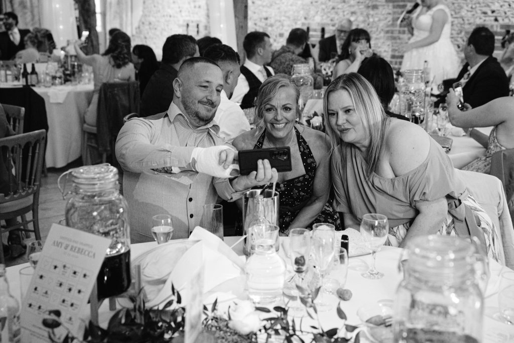 Three people seated at a table take a selfie together at a formal event, showcasing the lively atmosphere captured in Upwaltham Barns Wedding Photography, with other guests visible in the background.