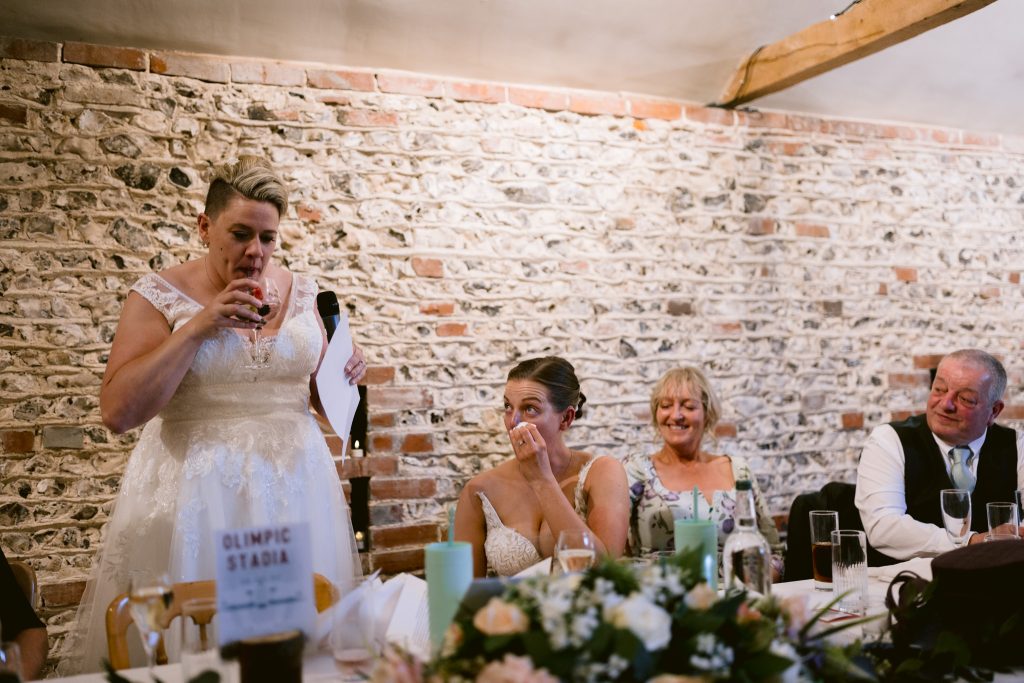 A bride in a white dress gives a speech at a table whilst another woman wipes away tears; two other guests smile and listen, capturing the emotion and joy of Upwaltham Barns Wedding Photography.