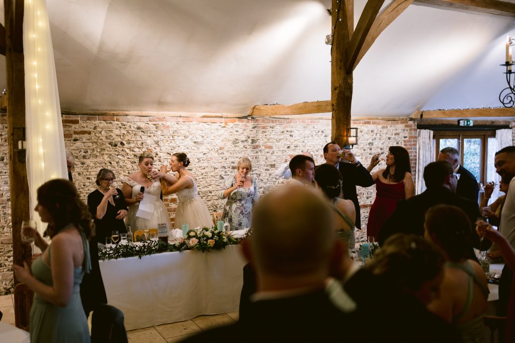 Wedding reception scene with guests raising glasses for a toast, two brides at the top table, and a rustic brick and wood interior—perfect inspiration for Upwaltham Barns Wedding Photography.