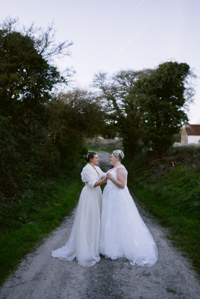 Two women in white wedding dresses stand on a rural path at Upwaltham Barns, holding hands and facing each other, surrounded by trees and greenery—capturing a beautiful moment of wedding photography.