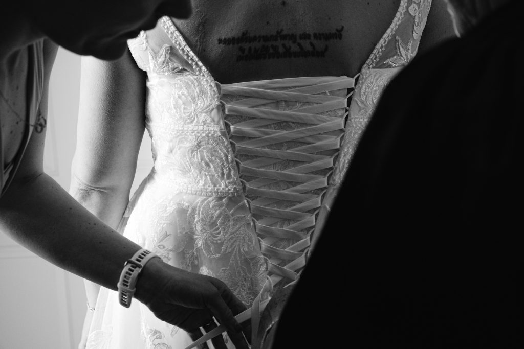 A person helps lace up the back of a wedding dress for another, revealing a tattoo on their upper back—a beautiful moment captured in timeless Upwaltham Barns Wedding Photography.