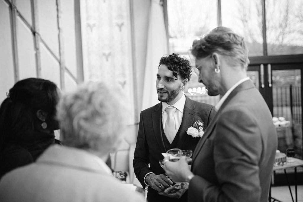 A man in a suit with a buttonhole smiles and chats with three guests at an indoor social event, holding a drink—captured in timeless Upwaltham Barns Wedding Photography style.