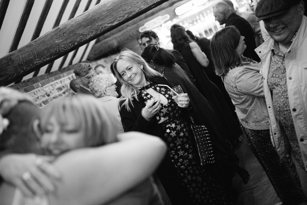 At an Upwaltham Barns wedding, a woman holding a drink stands in a crowded room, watching two people hug in the foreground whilst other guests mingle nearby.