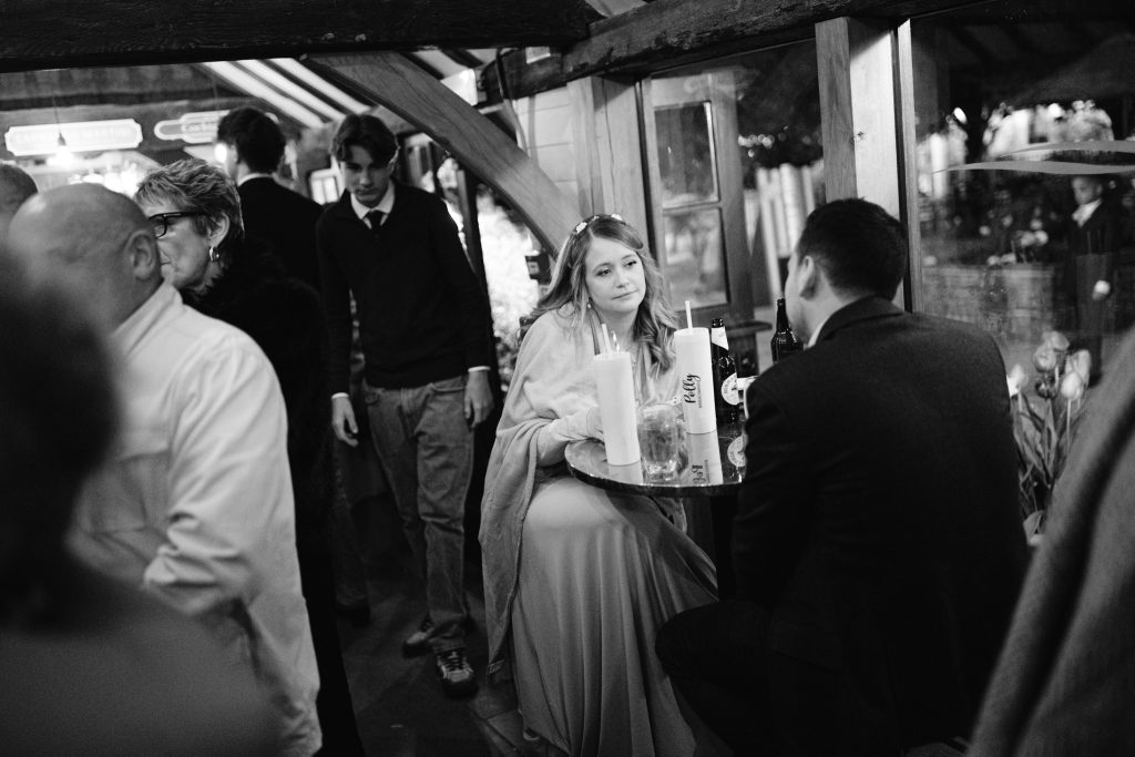 At a lively indoor gathering, a woman in a dress sits at a round table with drinks, gazing at a man opposite her—capturing the candid charm often seen in Upwaltham Barns Wedding Photography.