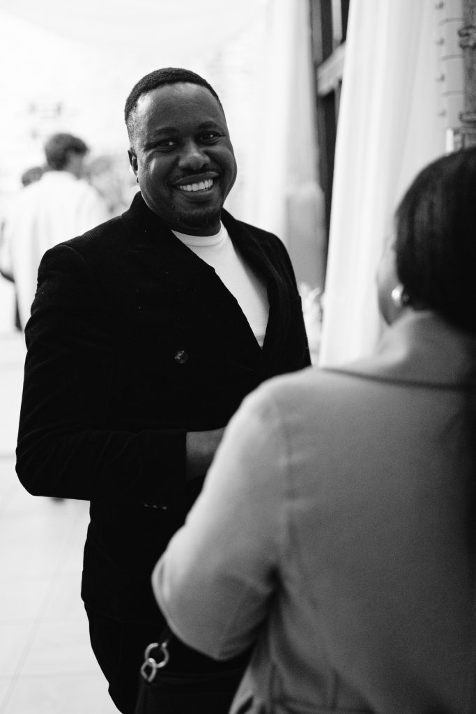 A man in a dark jacket smiles whilst speaking to a woman in a light coat at an indoor event. The black and white image captures the candid charm often seen in Upwaltham Barns Wedding Photography.