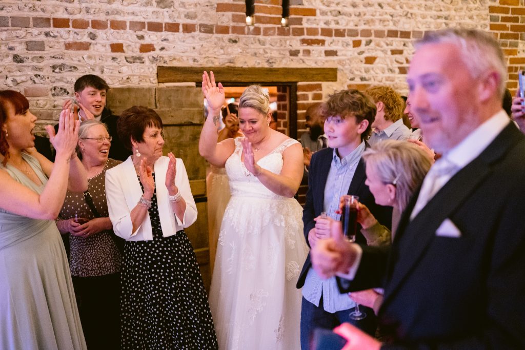 A bride in a white dress stands smiling and raising her arm, surrounded by guests clapping and celebrating in a warmly lit room with brick walls—perfectly capturing the joy of Upwaltham Barns Wedding Photography.