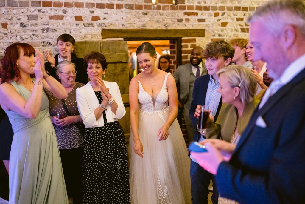 A bride in a white dress stands smiling among guests at an indoor Upwaltham Barns Wedding Photography session, with people applauding and chatting around her.