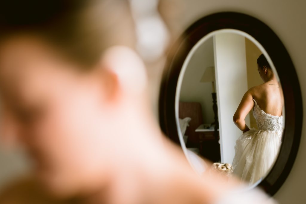 A woman in a wedding dress is seen from behind in the reflection of a round mirror, with her blurred profile in the foreground—capturing an intimate moment through Upwaltham Barns Wedding Photography.