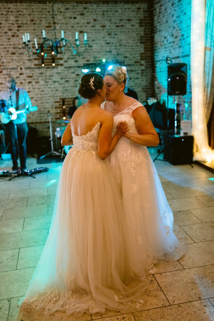 Two women in white wedding dresses share a dance and a kiss on the forehead in a warmly lit venue with exposed brick walls and a live musician—capturing the magic of Upwaltham Barns Wedding Photography.