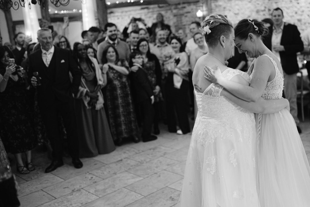 Two brides dance closely together in wedding dresses while a crowd of guests watches and smiles in the background, capturing a joyful moment of Upwaltham Barns Wedding Photography.