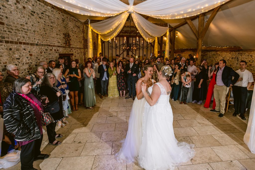 Two brides share a dance on the wedding dance floor, surrounded by guests in formal attire inside a beautifully decorated rustic venue—an unforgettable moment captured with Upwaltham Barns Wedding Photography.