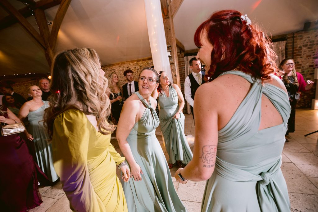 A group of people, including women in mint green dresses and one in yellow, dance and interact at an indoor event, with guests and fairy lights in the background—capturing the joyful moments of Upwaltham Barns Wedding Photography.