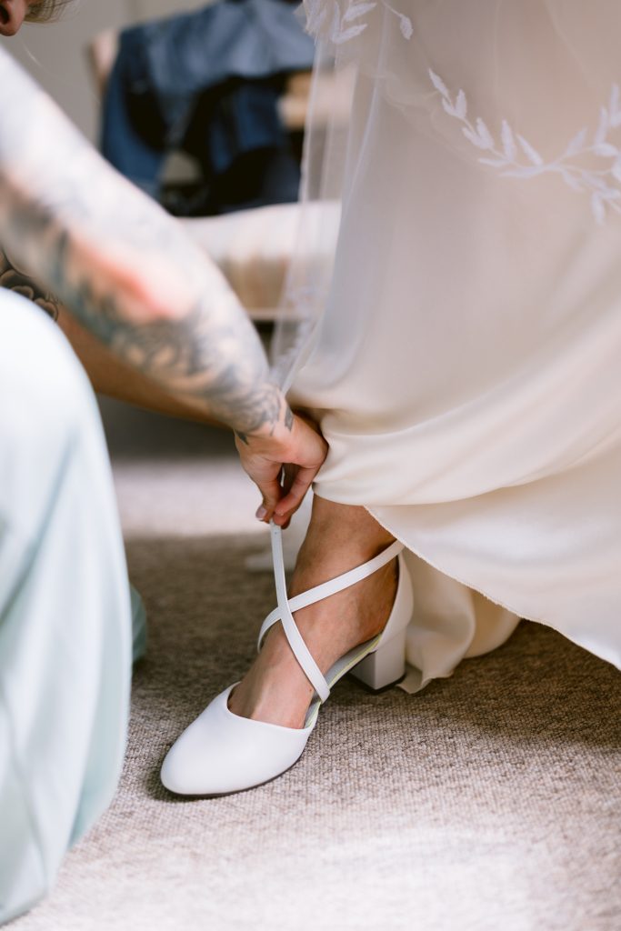 A person helps fasten the strap on a white shoe worn by someone in a white dress, seated on a carpeted floor—capturing an intimate moment typical of Upwaltham Barns Wedding Photography.