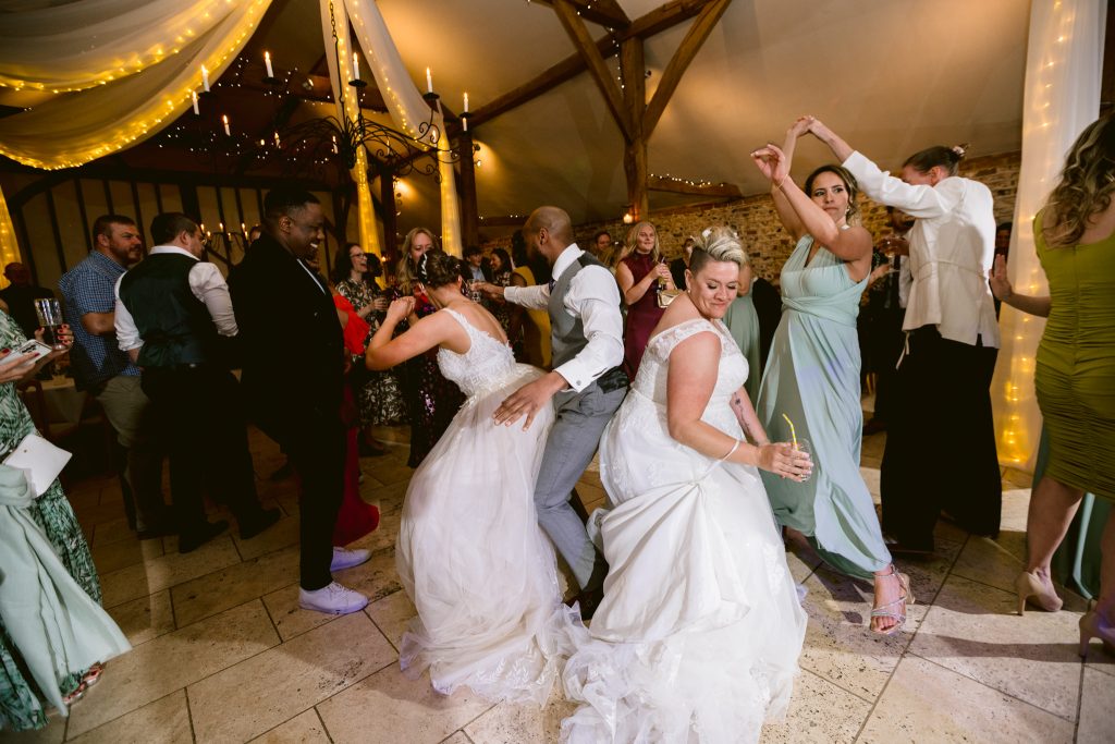 Wedding guests and newlyweds dance energetically on a decorated indoor reception floor at an Upwaltham Barns Wedding Photography event, surrounded by people in formal attire and festive lighting.