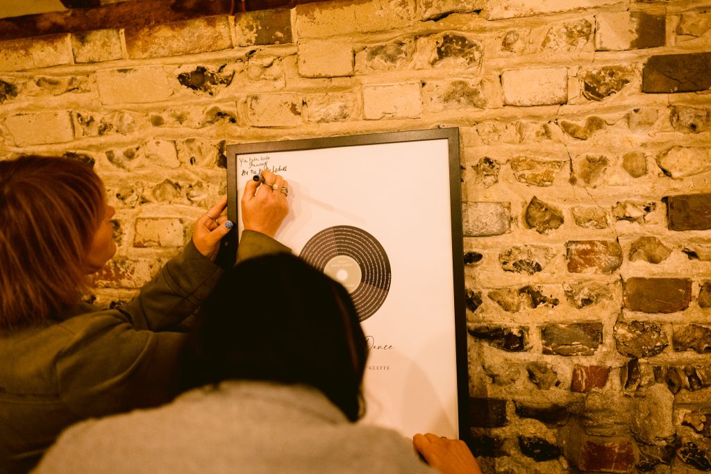 Two people write a message on a framed print featuring a graphic of a vinyl record, set against a brick wall—a charming moment captured during an Upwaltham Barns Wedding Photography session.