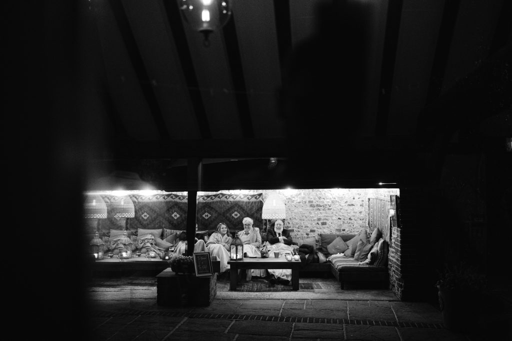 A group of people sit together in a cosy, covered outdoor seating area at night, illuminated by overhead lights—with patterned cushions and a brick wall in the background—capturing the warm ambience typical of Upwaltham Barns Wedding Photography.