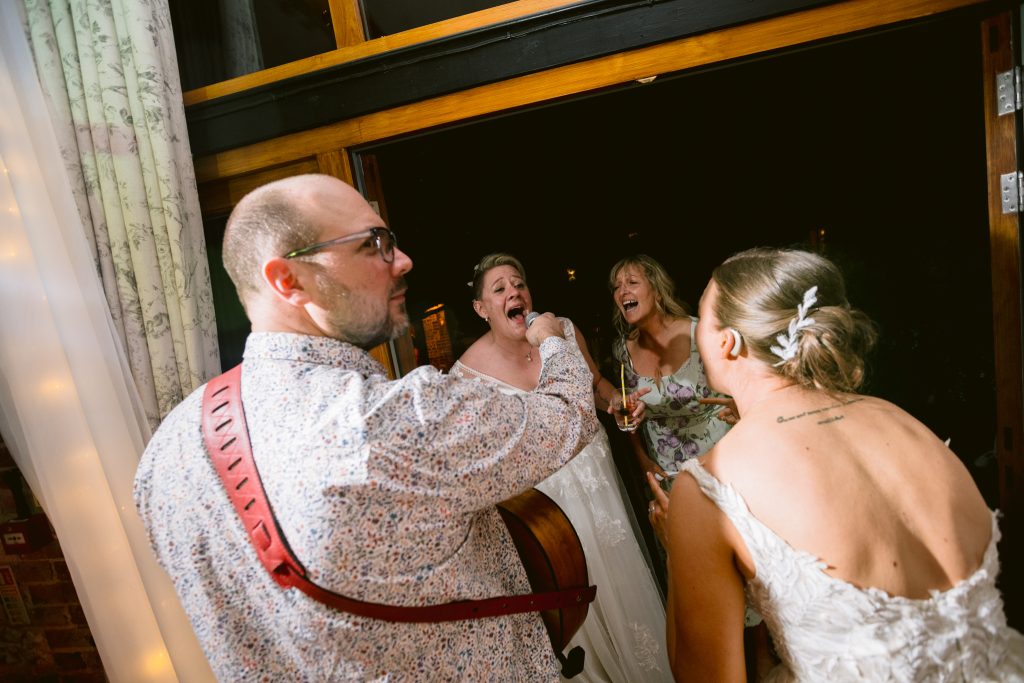 A man with a guitar plays music as three women, two in white dresses and one in a floral dress, sing and smile together indoors at a night event—capturing the joy of Upwaltham Barns Wedding Photography.