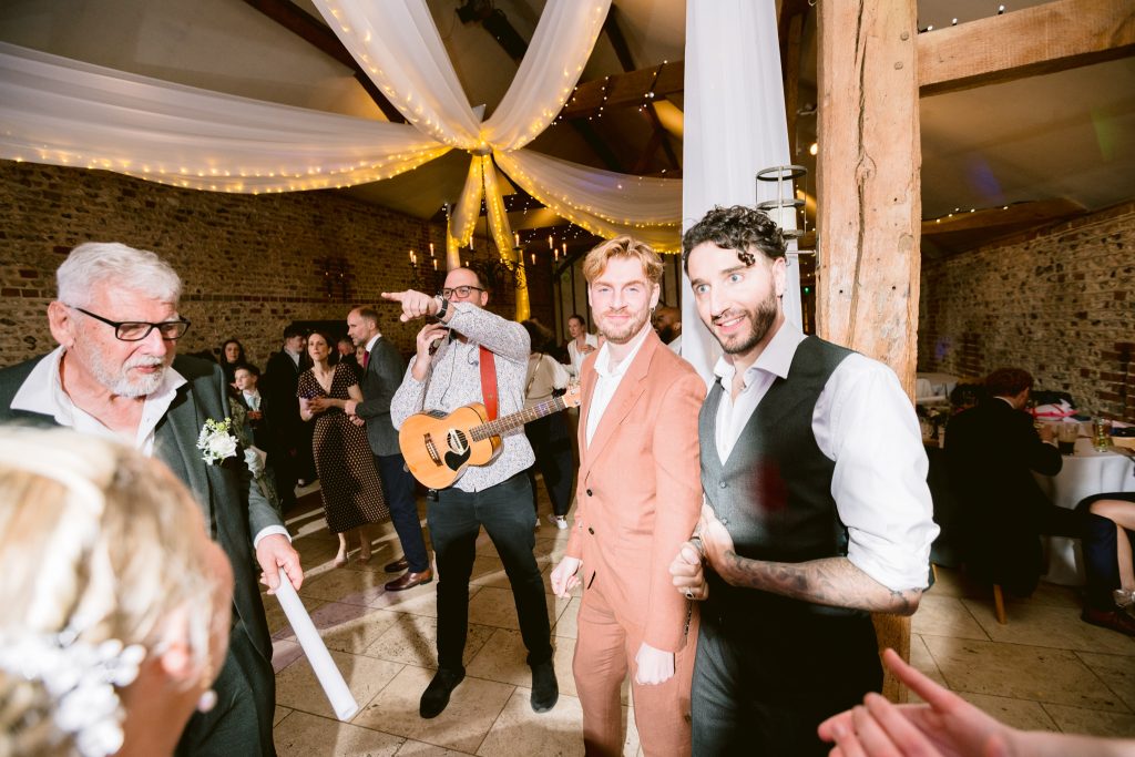 A group of people stand and smile at a wedding reception in a decorated venue with fairy lights and draped fabric; a musician with a guitar performs in the background, capturing the joy of Upwaltham Barns Wedding Photography.