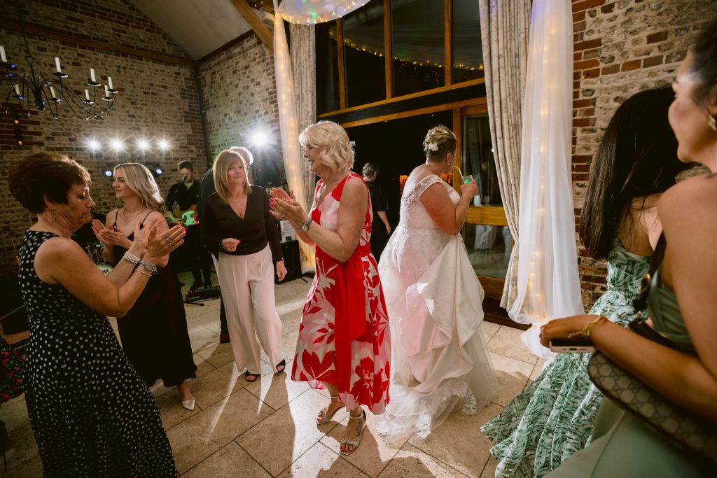 Several women, including a bride in a white dress, are dancing together at an indoor wedding reception with brick walls and fairy lights—a joyful moment captured for Upwaltham Barns Wedding Photography.