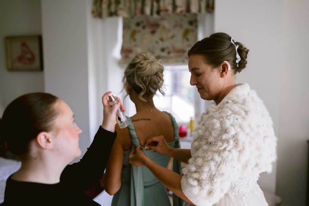 Three women stand in a room; one sprays a mist, another adjusts the dress of the third, who wears an off-white textured shawl. Capturing these moments is what makes Upwaltham Barns Wedding Photography so special. They are preparing for a formal occasion.