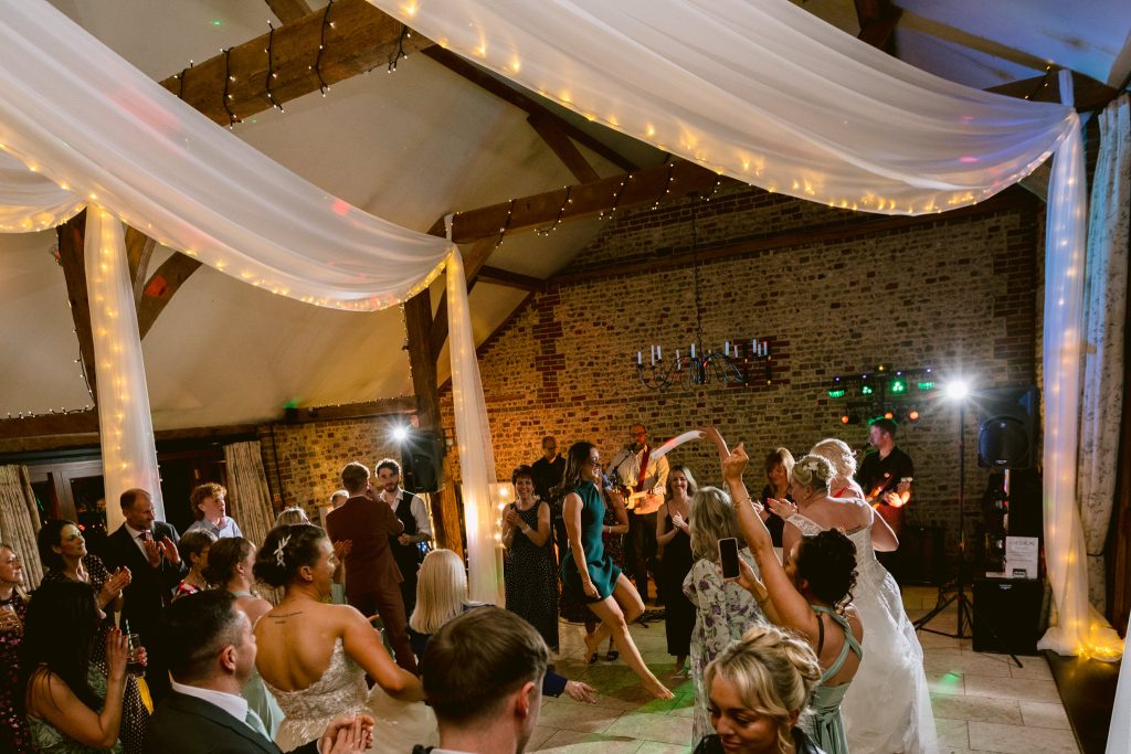 A group of people dance at a wedding reception in a decorated indoor venue with draped fabric, fairy lights, and a live band performing—capturing the joy of Upwaltham Barns Wedding Photography.