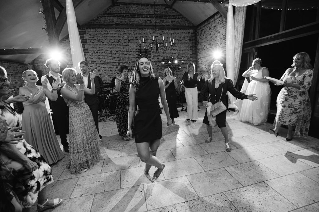 A group of people stand and clap as a woman dances in the centre of a well-lit hall with exposed brick walls and large windows, capturing the joy often seen in Upwaltham Barns Wedding Photography.