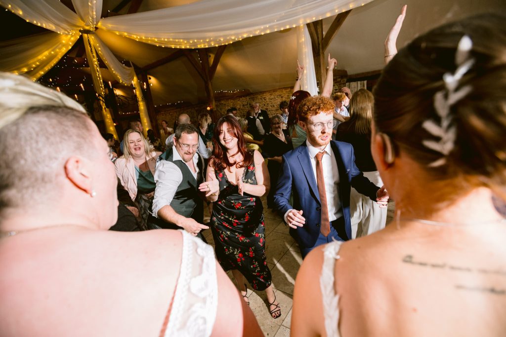 A group of people dance together at an indoor event with fairy lights and draped fabric overhead, capturing the joyous moments typical of Upwaltham Barns Wedding Photography.
