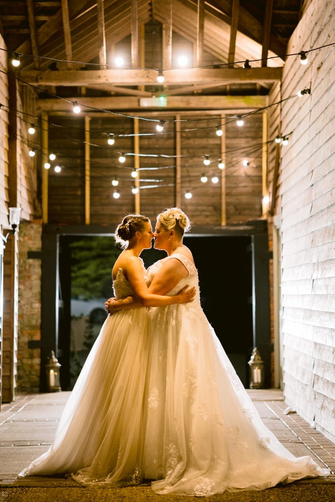Two brides in white wedding dresses stand face to face, embracing each other in a rustic, warmly lit barn decorated with fairy lights—capturing the magic of Upwaltham Barns Wedding Photography.