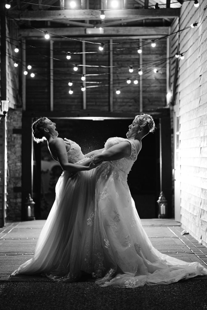 Two women in wedding dresses hold each other and laugh under fairy lights in a rustic barn, captured in classic black and white—an unforgettable moment of Upwaltham Barns Wedding Photography.