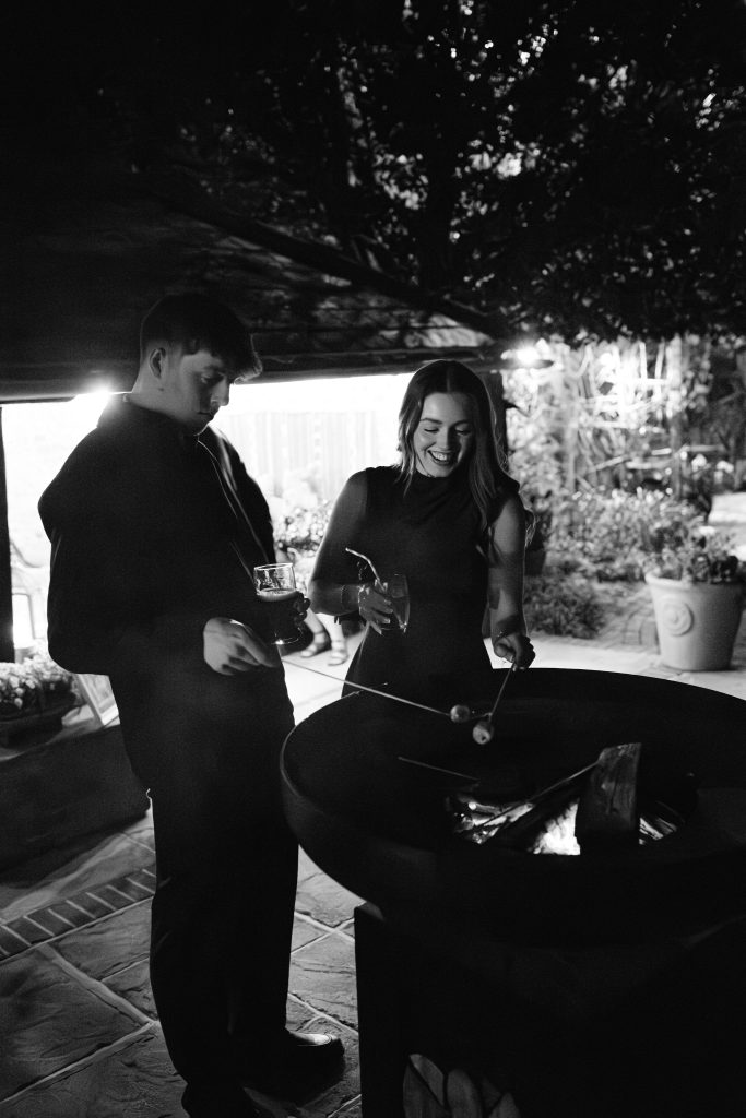 A man and woman stand by an outdoor firepit at night, toasting marshmallows on sticks and holding drinks—capturing the cosy atmosphere often found in Upwaltham Barns Wedding Photography.