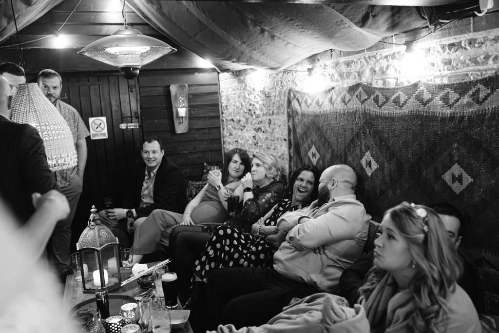 A group of people sit closely together on a sofa and chairs in a cosy, dimly lit room with patterned rugs on the wall, some holding drinks and talking—capturing candid moments perfect for Upwaltham Barns Wedding Photography.