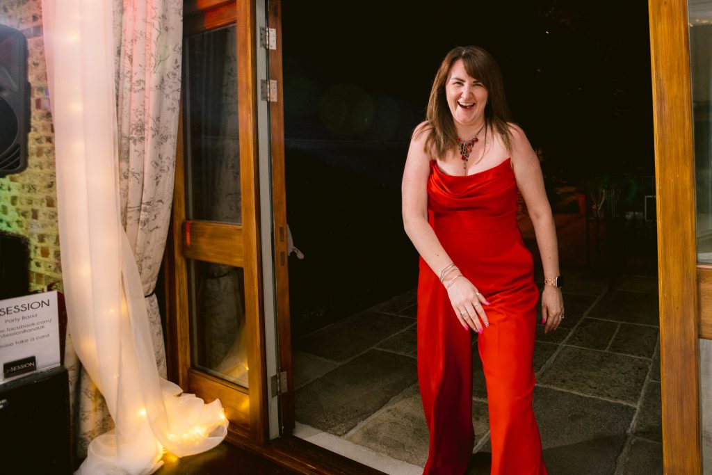 A woman in a red dress stands smiling in a doorway at night, with lights and curtains visible inside the room—capturing the warm ambience often sought after in Upwaltham Barns Wedding Photography.